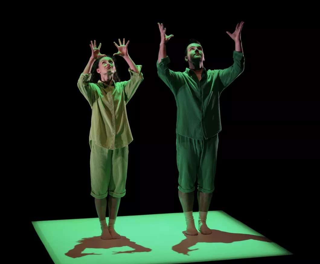 Two dancers in light green pajamas, arms raised above their heads, hands open, looking upward, standing on a green-lit platform casting their shadows against a black background.