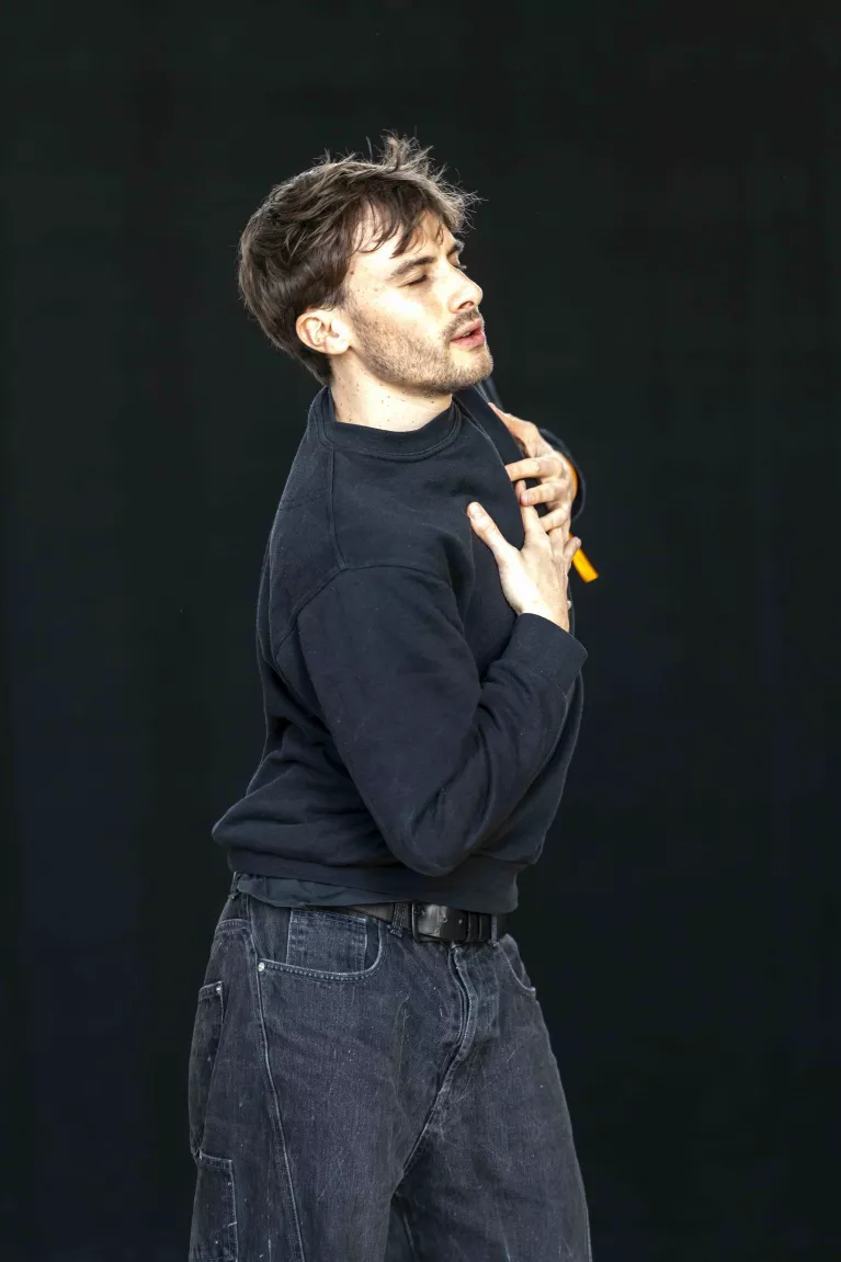 Un homme danse seule, habillé en noir, sur un fond noir. Il ramène ses mains vers son torse.