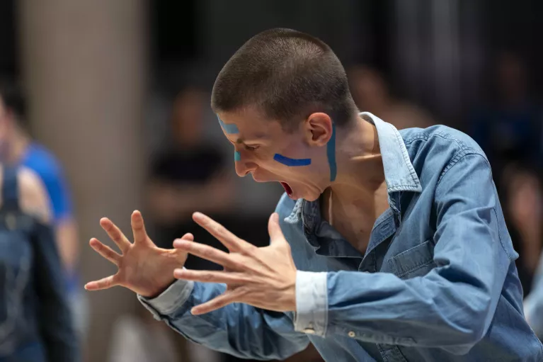 Un gros plan sur une personne qui semble hurler. Elle a de la peinture bleue sur le visage et est habillée en jean.