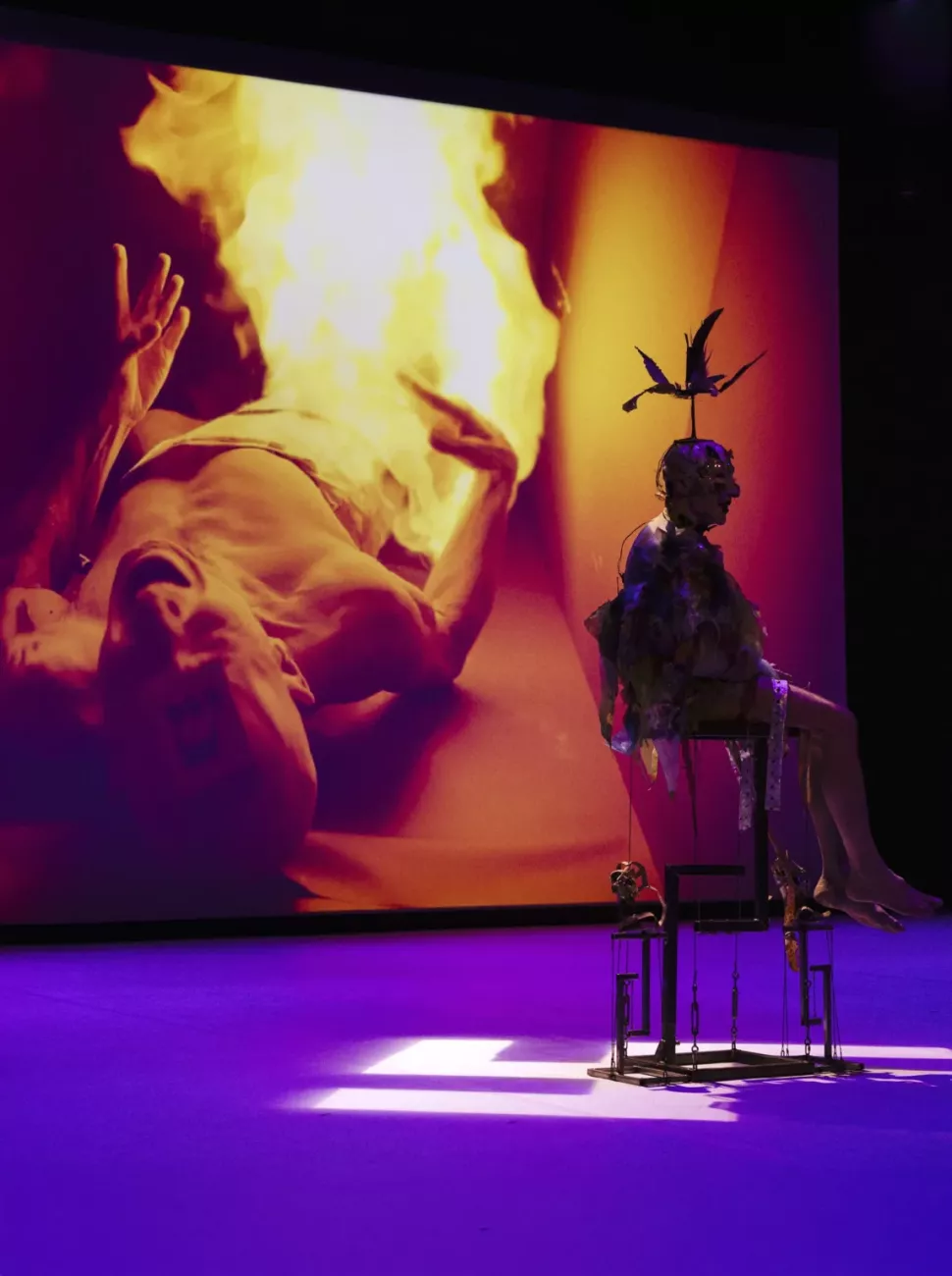 A figure in costume, wearing a bird-shaped headpiece, sitting on a metal structure on a purple stage, in front of a large screen projecting the image of a person on the ground surrounded by flames.