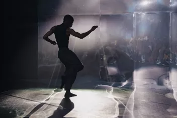 The silhouette of a dancer in mid-performance on stage, with one knee bent and one arm raised, starkly backlit. In the background, an audience can be glimpsed sitting in the reflection of a distorting mirror.
