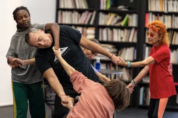Un groupe de personnes forme une chaîne en se tenant par les mains, dans une posture dynamique de mouvement collectif, dans une pièce avec des étagères de livres en arrière-plan.
