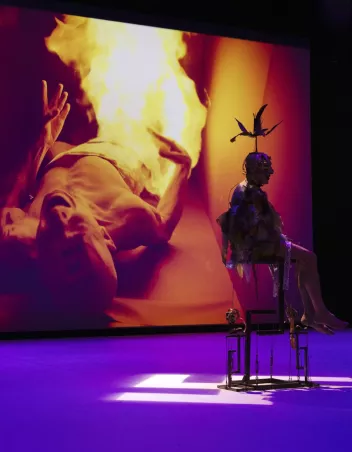 A figure in costume, wearing a bird-shaped headpiece, sitting on a metal structure on a purple stage, in front of a large screen projecting the image of a person on the ground surrounded by flames.