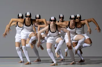 Un groupe de danseurs habillé de bonnets de bains et de lunettes de piscines se tiennent en formation sur une jambe avec les bras écartés
