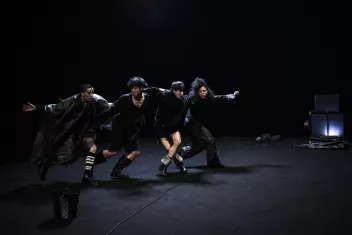 Four dancers dressed in black hold each other by the shoulders and move forward