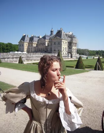 Une femme en costume d’époque fume une cigarette devant un château entouré de jardins à la française.