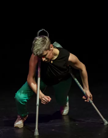 a photo of Claire Cunningham on stage, holding crutches, wearing a black vest top and green trousers
