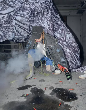A dancer kneeling on one knee, facing forward, is holding two cans of energy drink in front of some chillies on the floor
