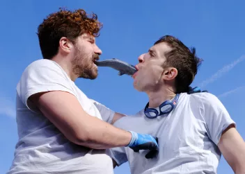 Photo of two men facing each other, each with a plastic fish in their mouth