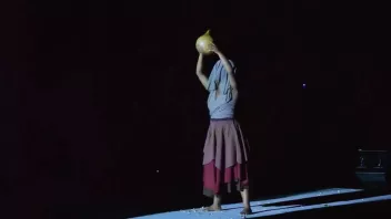 A dancer in a skirt, with a piece of fabric draped over her head, her arms raised as if holding a round object