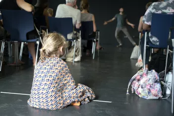 photo d'une enfant de dos, assise entre des chaise, qui regarde un spectacle 