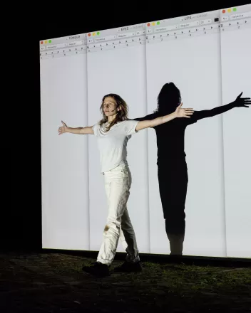 Photo du spectacle " Submission Submission"avec Bryana Fritz devant un écran qui projette des pages d'ordinateur, bras à l'horizontale
