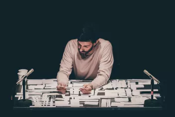 photo d'un spectacle avec un homme assis à une table qui contemple des bouts de papier