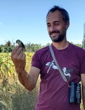 Photo of Maxime Zucca with a bird perched on his finger and binoculars around his neck