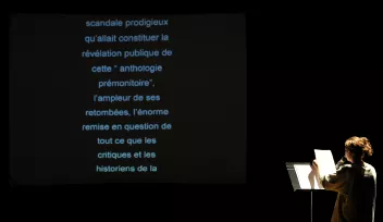 Une personne de dos lit un texte écrit sur une feuille face à un écran qui le retranscrit