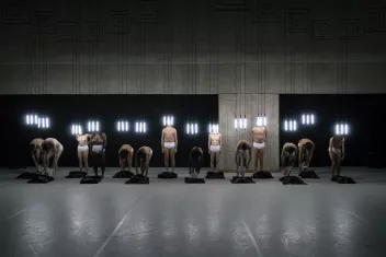 photo de danseurs en fond de scène vêtus de culottes blanches avec des tubes de lumières suspendus au dessus de la tête