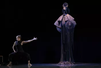 Une danseuse en position de seconde tend un bras vers une sculpture faite avec des habits