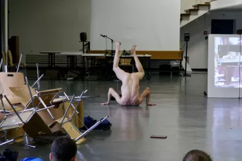 a performance by Xavier Le Roy, showing the naked artist from behind with one leg raised; on the left, a pile of chairs; on the right, a screen
