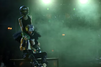 Two dancers wearing motorbike helmets – she’s in a tutu and he’s in motorbike trousers.