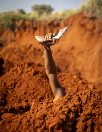 Photo of an arm emerging from the ground, holding a silver high-heeled shoe 
