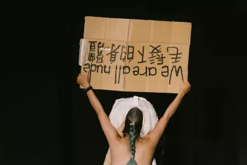 A dancer seen from behind holding a placard that reads ‘We are all nude’