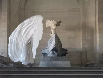 A dancer draped in a veil, like Loïe Fuller, standing next to a statue in the Louvre Museum