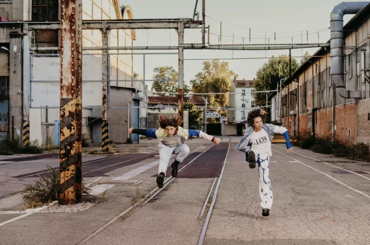 Deux danseuses sautent en l'air, bras écartés et cheveux en mouvement, sur une ancienne voie ferrée entre des bâtiments industriels désaffectés.
